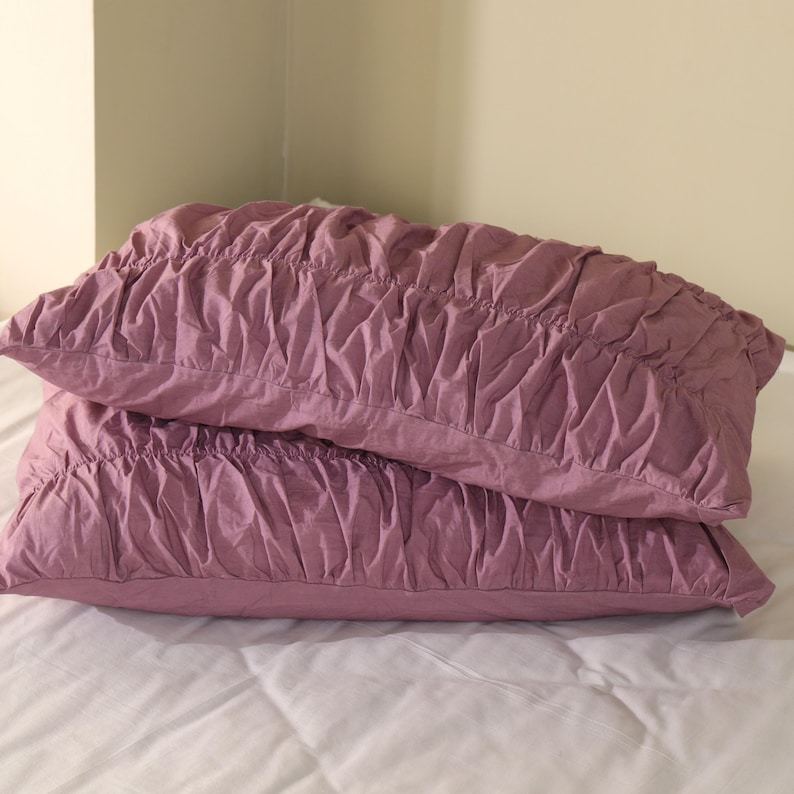 May include: Two mauve-colored decorative pillows stacked on a white bed. The pillows have a ruched, textured design across the top. The pillows are rectangular and appear to be made of a soft fabric.