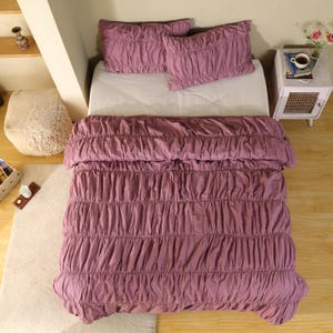 May include: A bed with a mauve ruched comforter and two matching pillowcases. The bedding is arranged on a white mattress. The room has a neutral color palette with a wooden floor and a small side table.