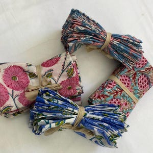 May include: Four bundles of patterned cloth napkins tied with twine. The napkins feature floral and fruit designs in pink, blue, and white. The bundles are arranged on a white surface.