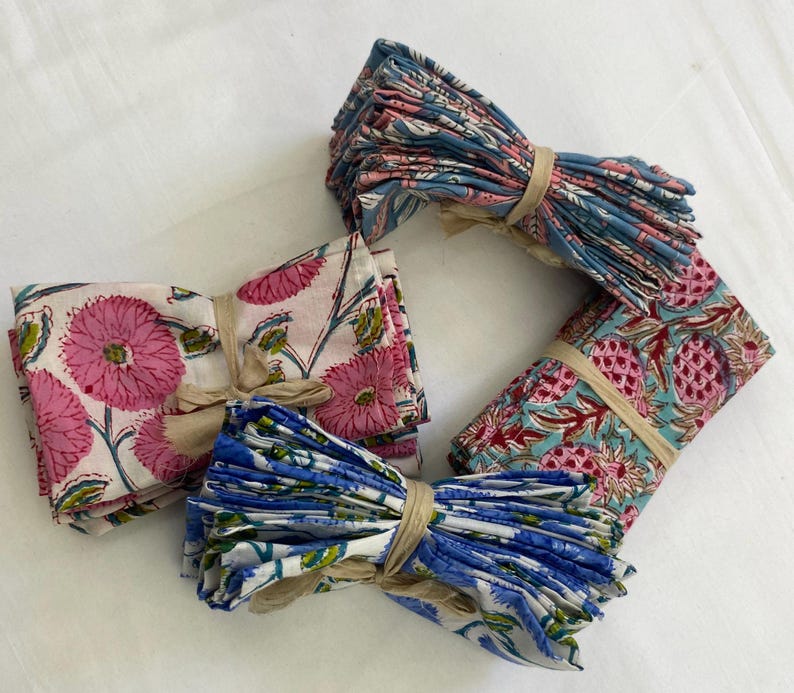 May include: Four bundles of patterned cloth napkins tied with twine. The napkins feature floral and fruit designs in pink, blue, and white. The bundles are arranged on a white surface.