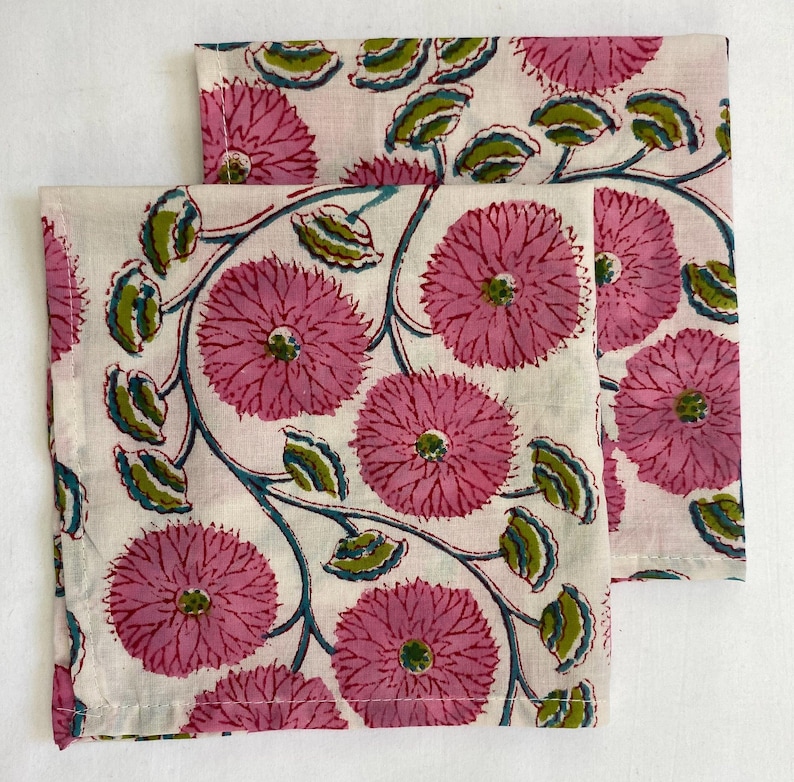 Op de afbeelding: Twee witte stoffen servetten met een herhalend bloemenpatroon. Het ontwerp toont grote roze bloemen, groene bladeren en turquoise ranken. De servetten zijn gevouwen en naast elkaar geplaatst, waardoor het volledige patroon wordt getoond.