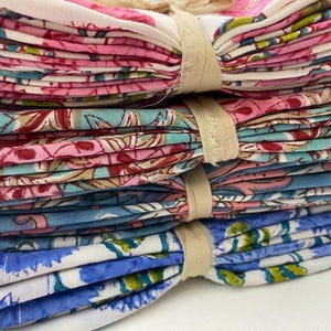 May include: A stack of folded floral patterned fabric napkins in pink, blue, and teal. The napkins are tied together with beige twine. The floral pattern includes flowers and leaves.