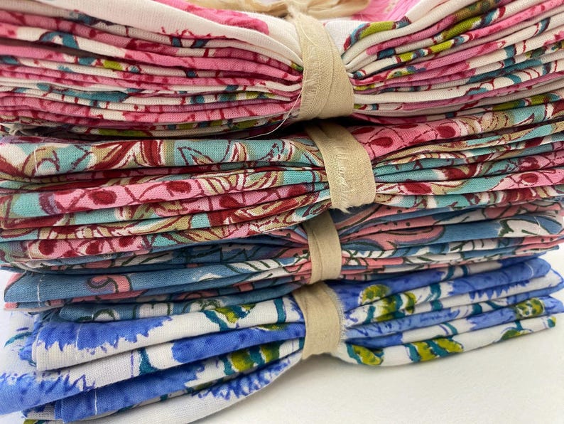 May include: A stack of folded floral patterned fabric napkins in pink, blue, and teal. The napkins are tied together with beige twine. The floral pattern includes flowers and leaves.