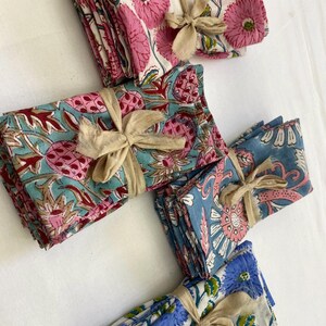 May include: A collection of folded floral print napkins in various colors, including pink, blue, and teal. Each stack is tied with a beige ribbon. The napkins feature detailed botanical designs.