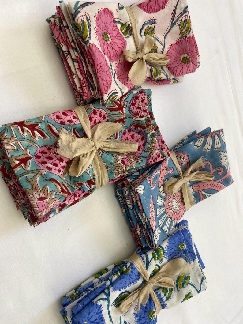 May include: A collection of folded floral print napkins in various colors, including pink, blue, and teal. Each stack is tied with a beige ribbon. The napkins feature detailed botanical designs.