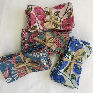 May include: Several stacks of floral print cloth napkins tied with beige ribbons. The napkins feature pink, blue, and teal floral patterns on a white background. The floral designs include large flowers and leaves. The napkins are neatly stacked.