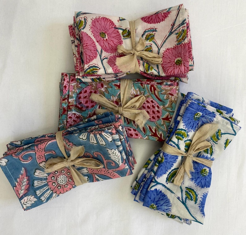 May include: Several stacks of floral print cloth napkins tied with beige ribbons. The napkins feature pink, blue, and teal floral patterns on a white background. The floral designs include large flowers and leaves. The napkins are neatly stacked.
