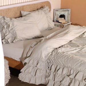 May include: A bed with light gray bedding, including a duvet cover and pillowcases with ruffled edges. The bed has a woven headboard and a wooden frame. The overall aesthetic is soft and inviting.