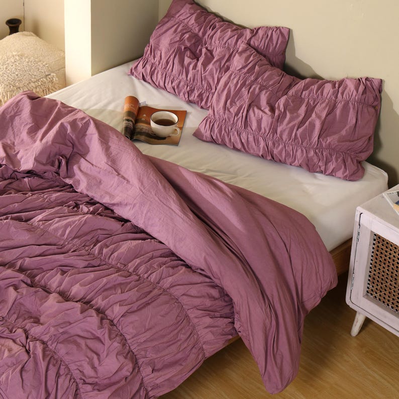 May include: A bed with a mauve comforter and two matching pillowcases. The bedding has a ruched design. A white sheet covers the mattress. A magazine and a cup of tea sit on the bed.