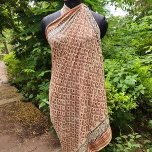 May include: A beige and brown patterned sarong displayed on a mannequin. The sarong features a repeating geometric design in shades of brown and red. The edges are trimmed with a decorative border in black, red, and yellow.