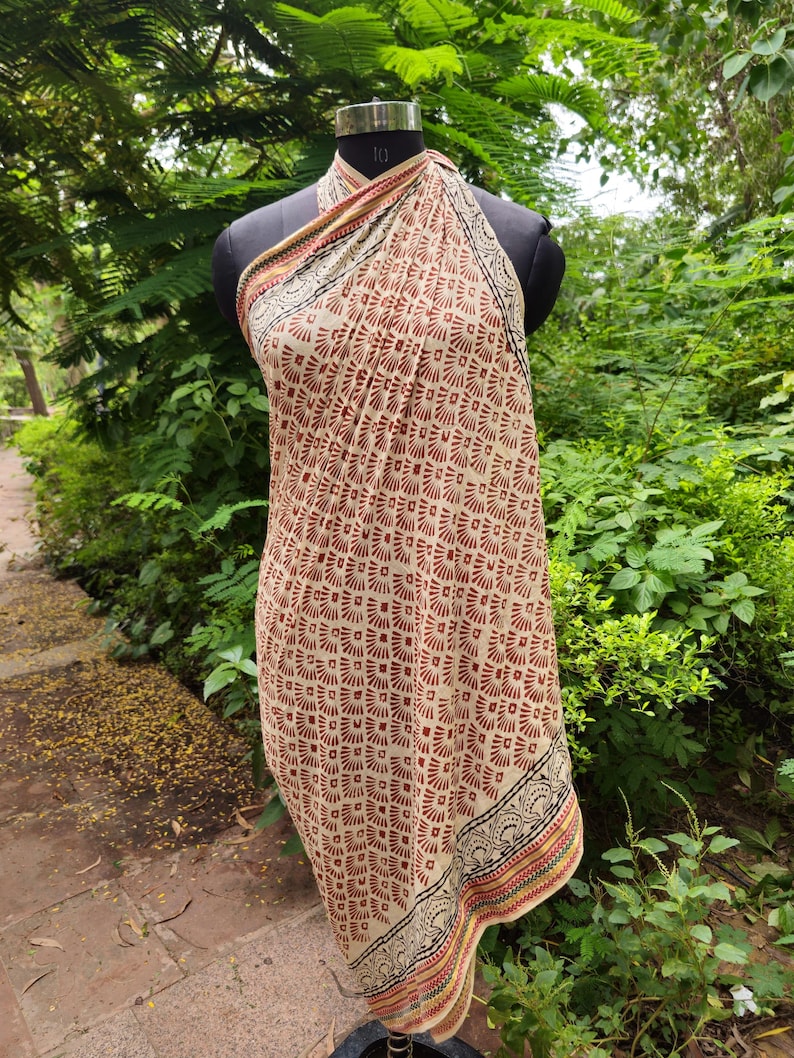 May include: A beige and brown patterned sarong displayed on a mannequin. The sarong features a repeating geometric design in shades of brown and red. The edges are trimmed with a decorative border in black, red, and yellow.