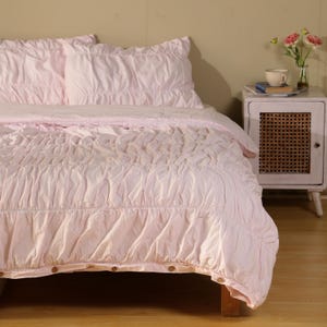 May include: A light pink bed set with a ruched design. The set includes a comforter and two pillowcases. The comforter has wooden buttons at the foot of the bed. A small white nightstand with a woven door is visible.