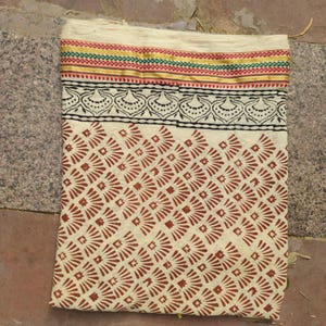 May include: Cream-colored textile with a repeating geometric pattern in reddish-brown. The top edge features a decorative border with red, green, and gold stripes, and a black and white floral design. The fabric is folded.
