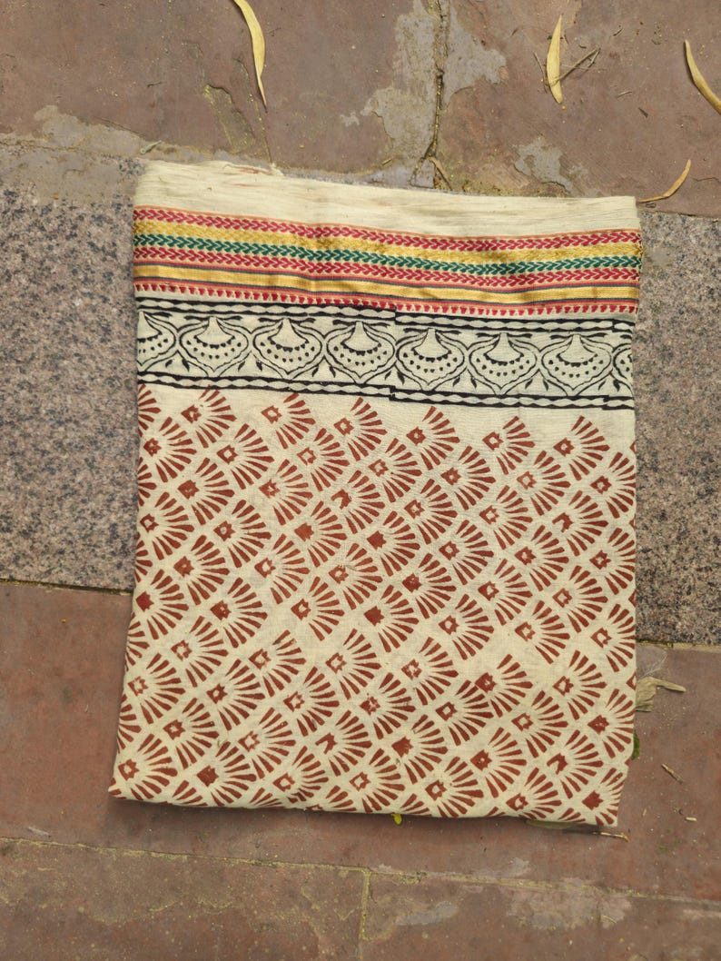 May include: Cream-colored textile with a repeating geometric pattern in reddish-brown. The top edge features a decorative border with red, green, and gold stripes, and a black and white floral design. The fabric is folded.