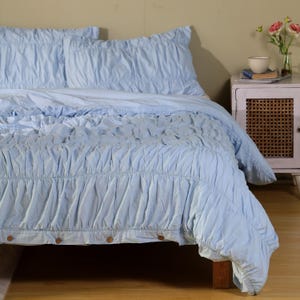 Puede incluir: Un juego de cama azul claro con un diseño con volantes. La colcha tiene una falda con botones de madera. La ropa de cama está sobre una estructura de cama de madera. Una mesita de noche con una puerta tejida y un jarrón de flores están en el fondo.