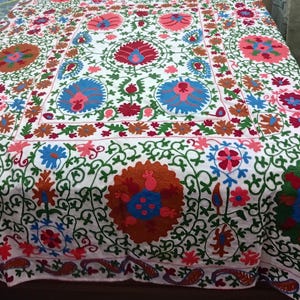 May include: White embroidered bedspread with a vibrant floral design. The bedspread features large, colorful flowers in shades of red, blue, orange, and pink, surrounded by green vines. The edges are trimmed with a detailed border.