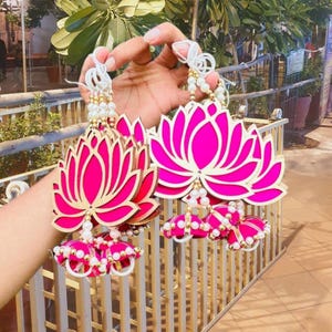 May include: Two decorative lotus flower ornaments. The flowers are hot pink with gold outlines and white and gold beaded accents. The ornaments are suspended from white cords and are held in a hand.