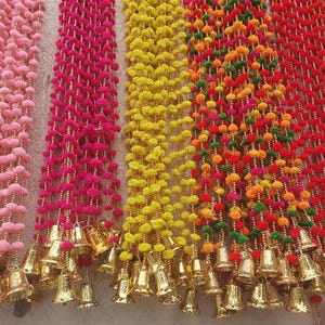 Puede incluir: Una colección de guirnaldas decorativas coloridas. Cada guirnalda presenta una cadena de pompones en tonos rosa, fucsia, amarillo, naranja, verde y rojo. Campanas doradas cuelgan en la parte inferior de cada guirnalda.
