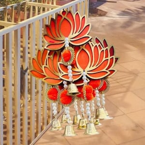 May include: A decorative wall hanging featuring three layered orange lotus flower cutouts with gold edges. The piece is adorned with multiple hanging strands of white faux pearls, bright orange pom-poms, and small metallic gold-toned bells.