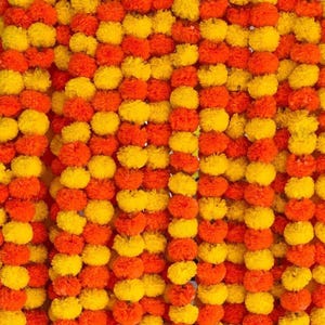 Op de afbeelding: Een dichte verticale opstelling van kunstmatige goudsbloem-slingers met afwisselende strengen van feloranje en heldergele pompon-achtige bloesems. De bloemen zitten dicht op elkaar gepakt om een vol, getextureerd gordijn te vormen.