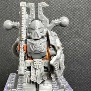 May include: A gray miniature figure of a warrior in armor, holding a weapon. The figure has a helmet with horns and a detailed chest plate. The miniature is on a small base, with orange and yellow accents.