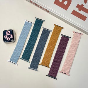 Puede incluir: Correas trenzadas para relojes inteligentes en varios colores: azul claro, verde azulado, gris, amarillo mostaza, burdeos y rosa. Un reloj inteligente muestra la hora. El texto "Bella Italia" es visible.