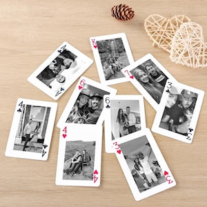 Puede incluir: Un conjunto de cartas de juego con fotografías en blanco y negro. Las cartas están dispersas sobre una superficie de madera clara, con una piña y un corazón decorativo en el fondo. Cada carta muestra una foto diferente de personas.