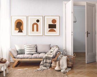 May include: Three framed abstract art prints with geometric shapes in brown and beige tones hang above a light wood sofa with pillows and a striped throw. A woven rug and a small side table with a basket complete the room's decor. The room has white walls and a wooden floor.