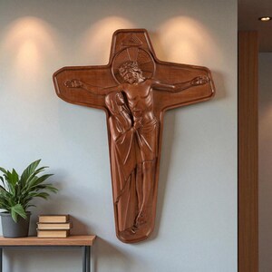 May include: A large, intricately carved wooden crucifix depicting Jesus Christ on the cross, with a figure at the base. The sculpture is a warm brown color and mounted on a light blue wall. The cross has a unique shape with rounded edges.