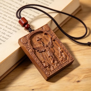 May include: A rectangular wooden pendant featuring a detailed relief carving of a winged figure holding a cross, suspended from a dark brown cord with a matching wooden bead. The border is decorated with small circular motifs and intricate patterns.