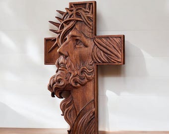 May include: A wooden cross featuring a detailed relief carving of a man's face, wearing a crown of thorns. The cross is a dark brown color and is displayed on a wooden surface.