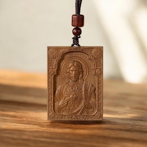 Pode incluir: Pingente retangular de madeira com uma imagem esculpida de Jesus com uma auréola, com a mão no coração. Pendurado por um cordão marrom com contas de madeira. O pingente é de cor marrom quente.