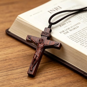 Könnte beinhalten: Ein dunkelbraunes Holzkruzifix mit einer geschnitzten Jesusfigur und den Buchstaben INRI an der Spitze. Das Kreuz ist an einer schwarzen Kordel befestigt und liegt auf einem aufgeschlagenen Buch mit sichtbarem Text, darunter John 3:16.