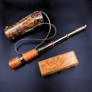 Puede incluir: Un telescopio antiguo de latón y cuero con una caja de madera y un estuche de cuero decorativo. El telescopio tiene un agarre de cuero y un cuerpo de metal plateado. El estuche es marrón con diseños ornamentados y una correa. La caja de madera es rectangular.