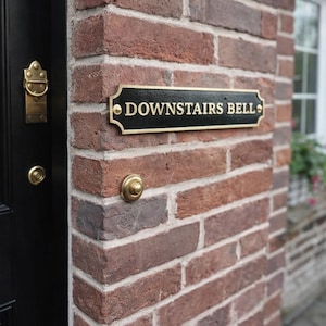 Può includere: Un cartello rettangolare nero e dorato con la scritta "DOWNSTAIRS BELL" è montato su un muro di mattoni rossi. Il testo e la cornice sono dorati. Sotto c'è un pulsante del campanello dorato. A sinistra c'è una porta nera.
