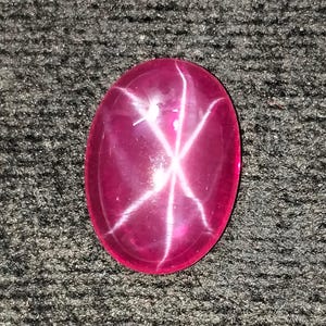 Könnte beinhalten: Ovale, tiefrote Edelstein mit einem sechsstrahligen Sternmuster. Die polierte Oberfläche reflektiert das Licht und erzeugt einen Sterneffekt. Der Edelstein liegt auf einem dunklen, strukturierten Hintergrund.
