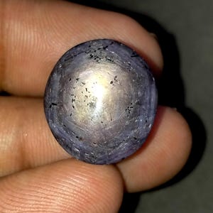 Könnte beinhalten: Ein runder, im Cabochon-Schliff gefertigter Sternsaphir-Edelstein mit einem durchscheinenden, gräulich-violetten Farbton. Der Stein zeigt einen ausgeprägten sechsstrahligen Sterneffekt in der Mitte sowie sichtbare dunkle Einschlüsse und Texturen.