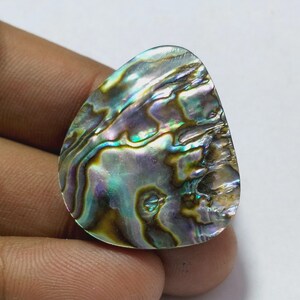 Könnte beinhalten: Eine tropfenförmige Abalone-Schale mit schillernden Farben wie Grün, Blau, Lila und Braun. Die polierte Oberfläche reflektiert das Licht und zeigt die natürlichen Muster und Texturen der Schale. Die Schale wird in einer Hand gehalten.