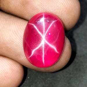 Könnte beinhalten: Oval geschliffener Rubin-Edelstein mit einem sechsstrahligen Sternmuster. Der Edelstein hat eine leuchtend rote Farbe mit einem hellen, weißen Sterneffekt auf seiner Oberfläche. Der Stein ist glatt und gut geschliffen, was seine natürliche Schönheit zeigt.