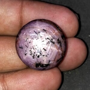 Könnte beinhalten: Ein runder, polierter Edelstein mit einem hellvioletten Farbton. Der Stein hat schwarze Einschlüsse und eine reflektierende Oberfläche. Der Edelstein wird in einer Hand gehalten, um seine Größe und Form zu zeigen. Das Gesamtbild deutet auf eine natürliche, unraffinierte Ästhetik hin.