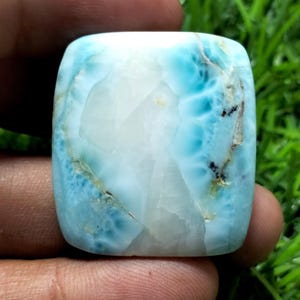 Puede incluir: Una gema pulida de corte cuadrado con apariencia veteada. La piedra presenta una mezcla de tonos blancos y azul claro, con vetas azules más oscuras e inclusiones marrones. La gema se sostiene en una mano, con un fondo verde.