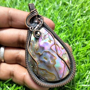 Könnte beinhalten: Tropfenförmiger Anhänger mit einem kupferdrahtumwickelten Rahmen. Der Anhänger zeigt eine irisierende Abalone-Schale mit violetten, grünen und goldenen Farbtönen. Die Drahtarbeit ist aufwendig und bildet eine dekorative Bordüre und eine Öse für eine Halskette.