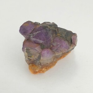 Könnte beinhalten: Ein rohes Mineralexemplar mit mehreren durchscheinenden, violetten Amethystkristallspitzen, die zusammen gruppiert sind. Die Basis des Clusters zeigt eine texturierte, erdig orange-braune Matrix, während die oberen Bereiche tiefe Violetttöne aufweisen.