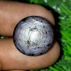Könnte beinhalten: Ein runder, im Cabochon-Schliff gefertigter Sternsaphir-Edelstein mit gräulich-violetter Färbung. Der Stein zeigt einen ausgeprägten, sechsstrahligen Sterneffekt in der Mitte sowie sichtbare dunkle Einschlüsse und eine glatte, polierte Oberfläche.