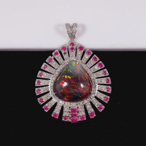 Könnte beinhalten: Tropfenförmiger Anhänger mit einem großen, dunklen Opal, der lebendige Farben zeigt. Der Opal ist von kleinen, roten Rubinen und funkelnden weißen Steinen umgeben, gefasst in einem silberfarbenen Metall.