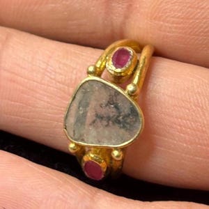 Könnte beinhalten: Ein Goldring mit einem großen, unregelmäßigen, durchscheinenden grauen Stein mit dunklen dendritischen Einschlüssen in der Mitte, flankiert von zwei kleinen, ovalen, tiefrosa Rubinen in Goldfassungen auf einer geteilten Ringschiene.