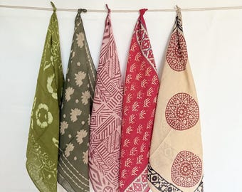 May include: Five patterned bandanas in shades of green, pink, and red hang from a clothesline against a white background. Each bandana features a unique design, including floral, geometric, and circular patterns. The bandanas are made of a lightweight fabric.