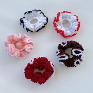 May include: Five handmade crochet scrunchies in various colors: white with black trim, white with red trim, light pink with white trim, dark red with white trim, and brown with white trim. These hair accessories are perfect for adding a touch of handmade charm to any hairstyle.