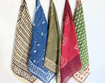 May include: Five colorful bandanas hanging from a string against a white background. The bandanas feature various patterns and colors, including beige with black lines, blue with white designs, olive green with white floral patterns, red with black geometric patterns, and green with white paisley.
