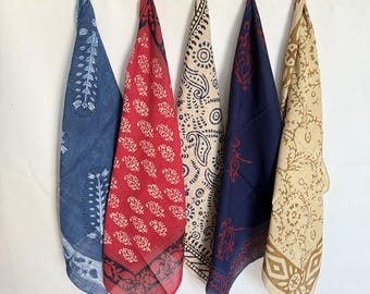 May include: Five patterned bandanas hanging from a string against a white background. The bandanas feature various designs in shades of blue, red, beige, and navy. Each bandana is knotted at the top, showcasing the fabric's patterns and colors.